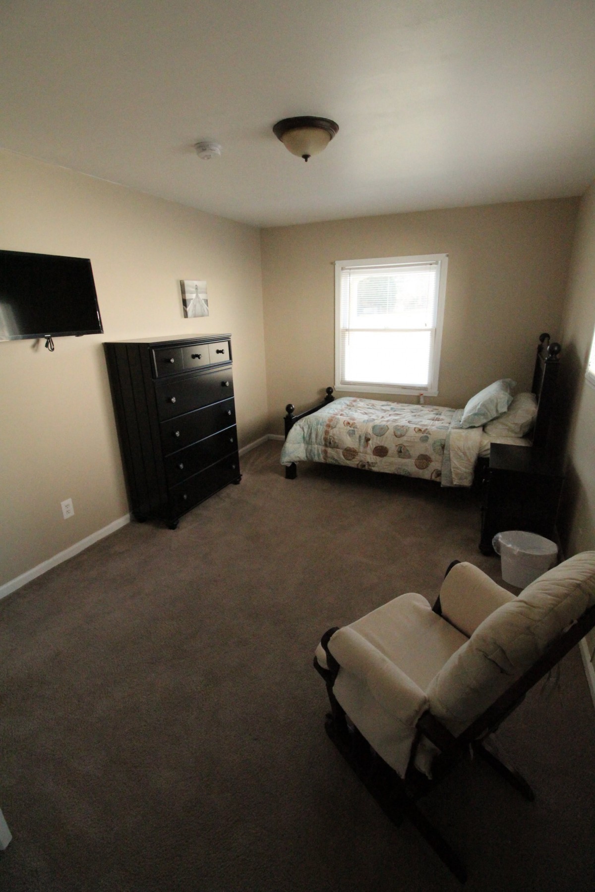 A tidy bedroom with a bed, dresser, mounted TV, and a window.