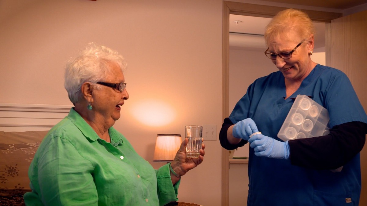 An elderly woman with a glass of water receives medication from a nurse.