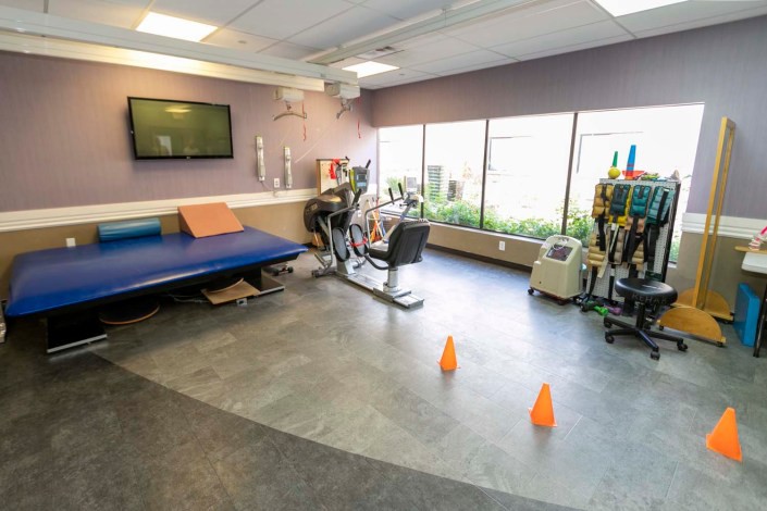 A rehabilitation room equipped with exercise equipment, including a bicycle machine and treatment table, with cones on the floor.