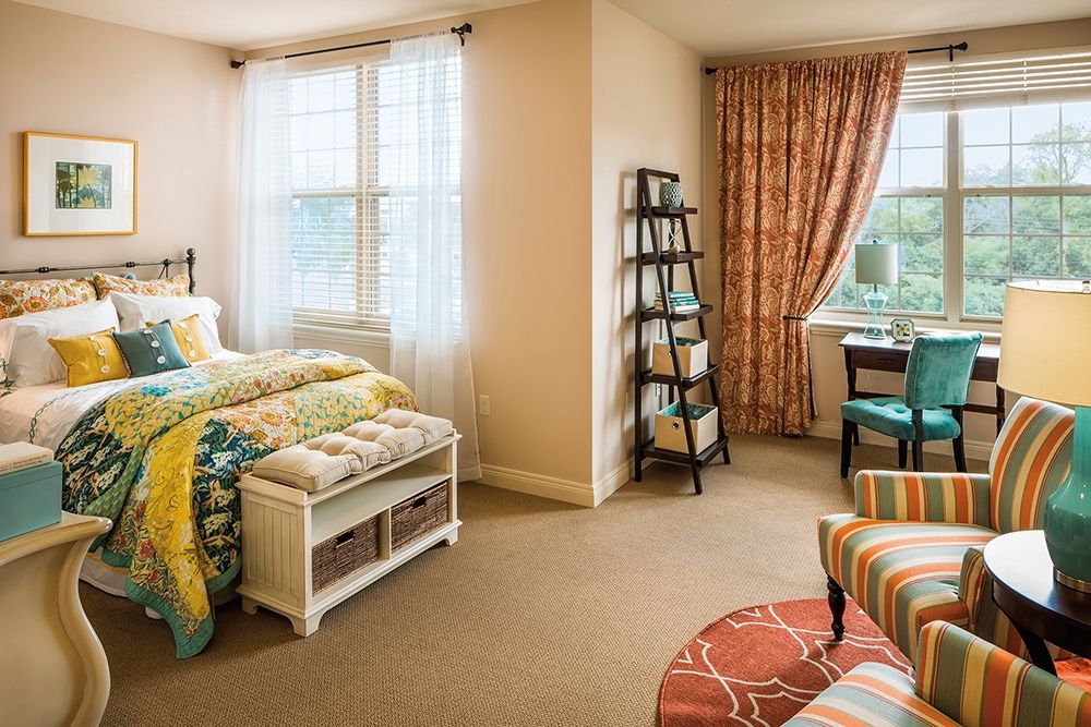 A cozy bedroom with colorful bedding, a small bookshelf, and natural light from large windows.