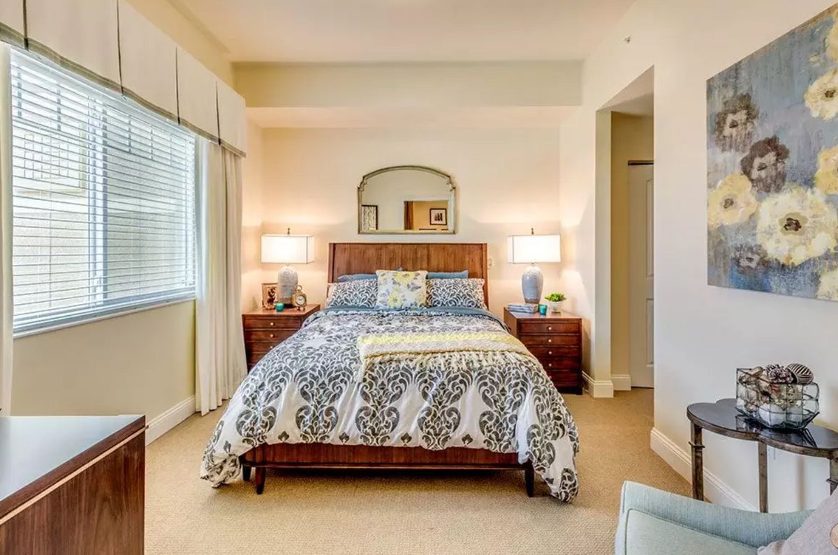 A cozy bedroom with a neatly arranged bed, flanked by two bedside tables and lamps, featuring a decorative mirror above the headboard.