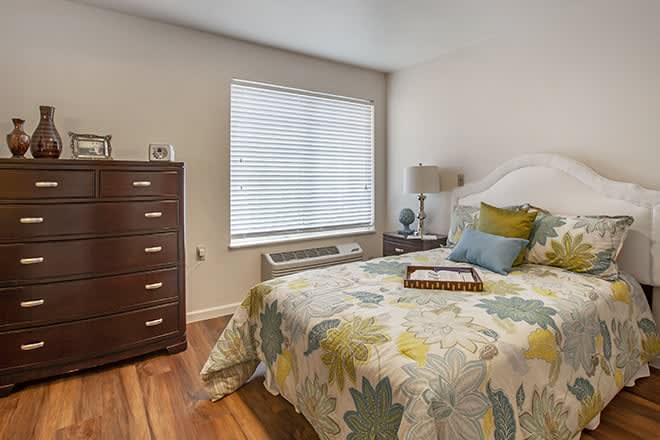 A cozy bedroom with a floral bedspread, wooden dresser, and window with blinds.