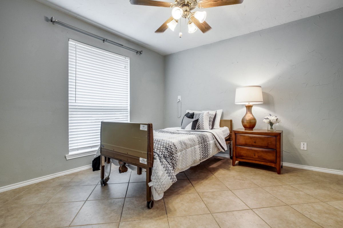 A small bedroom with a hospital bed, nightstand, and lamp under a ceiling fan.