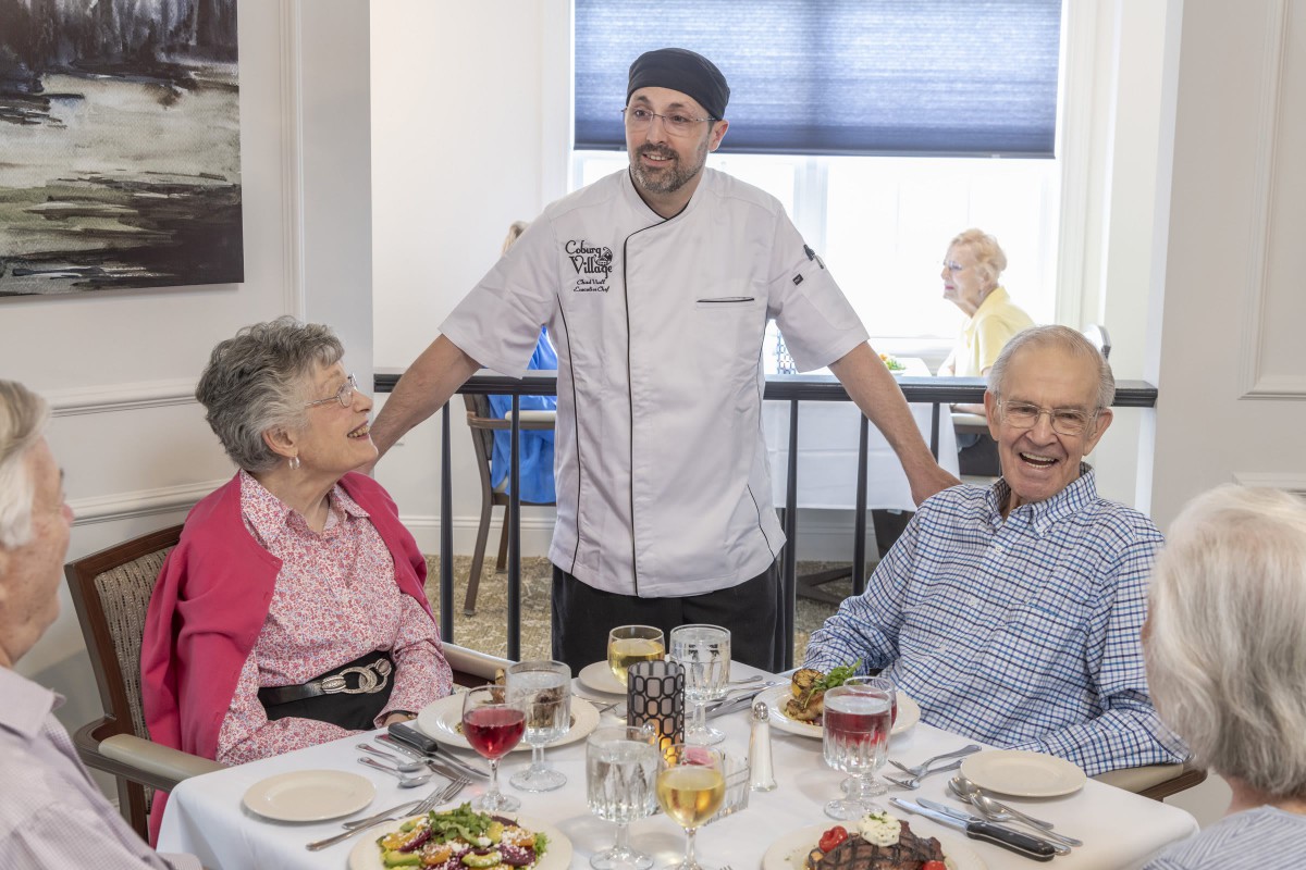 A chef standing with two senior patrons enjoying a meal at a restaurant.
