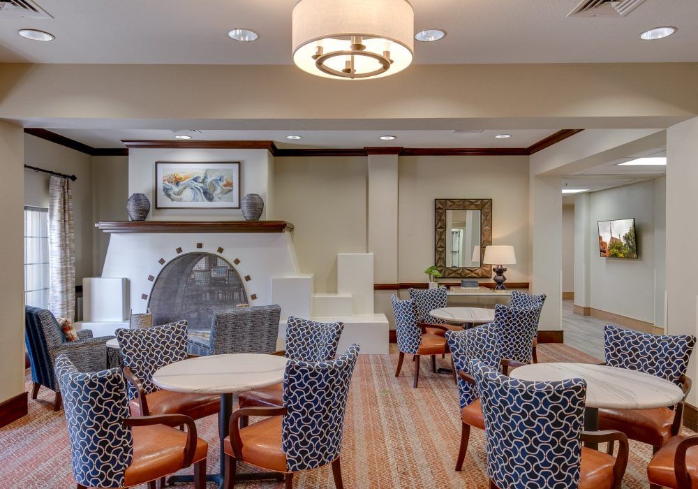 A modern seating area with patterned chairs and tables arranged around a central fireplace, illuminated by stylish lighting.