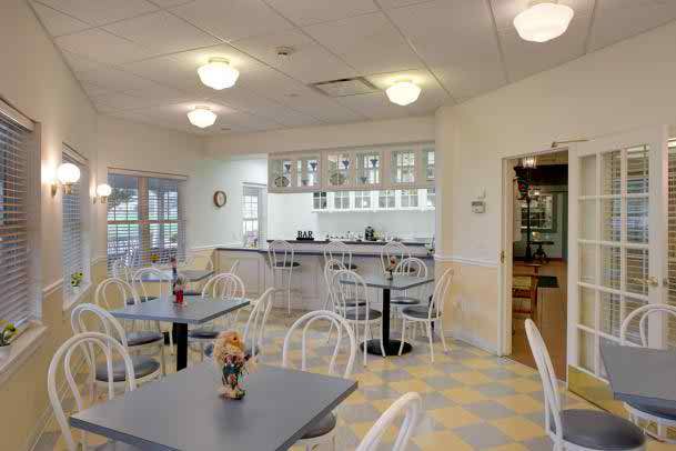 A bright and tidy cafeteria with multiple tables and chairs arranged neatly, featuring ceiling lights and a tiled floor.