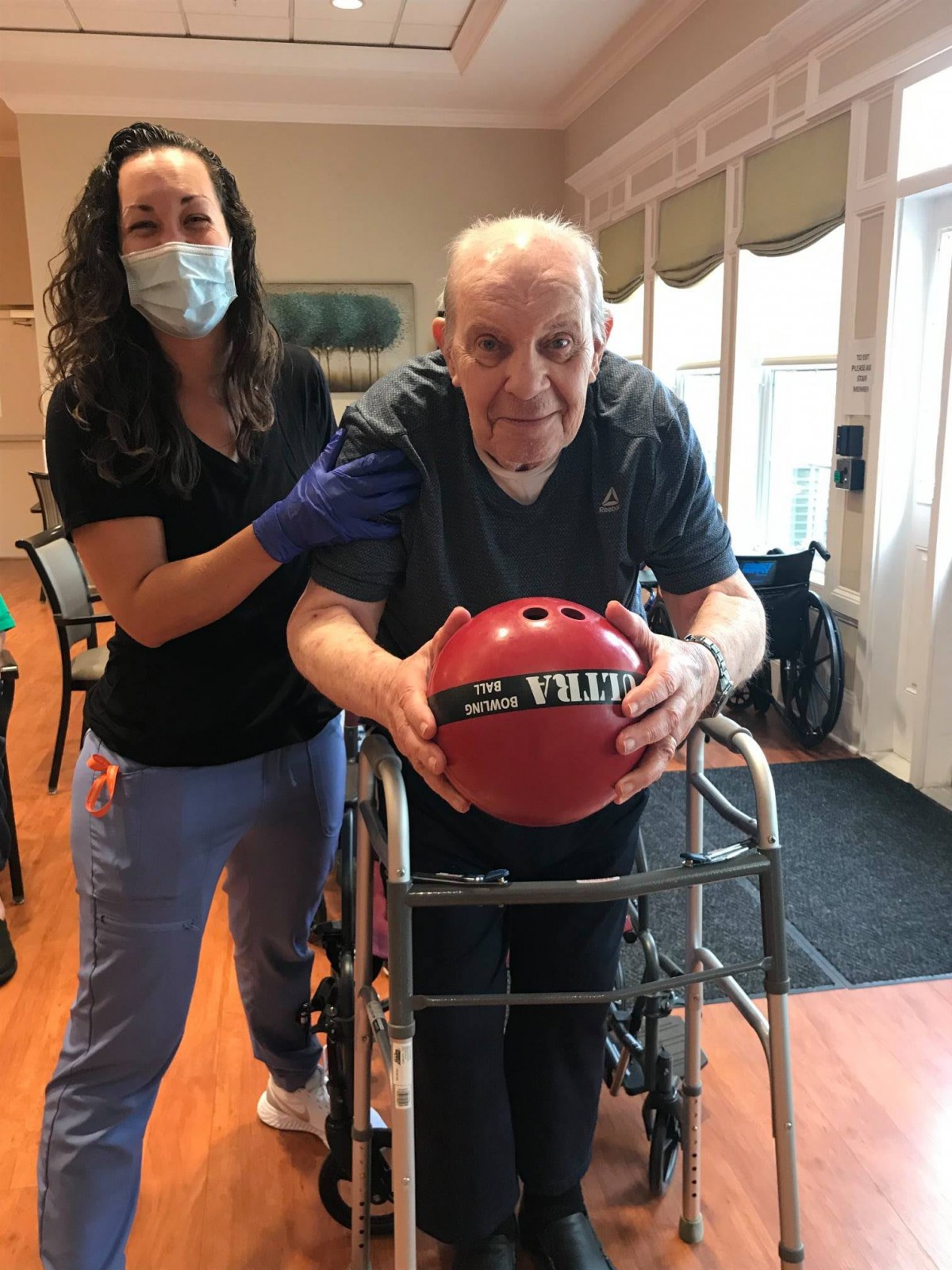 An elderly man with a walker is assisted by a caregiver as he holds a bowling ball indoors.