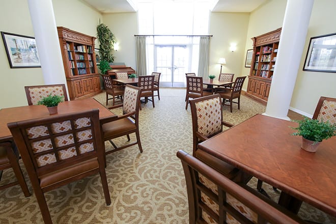 A well-lit library interior with wooden tables and chairs, surrounded by bookshelves.
