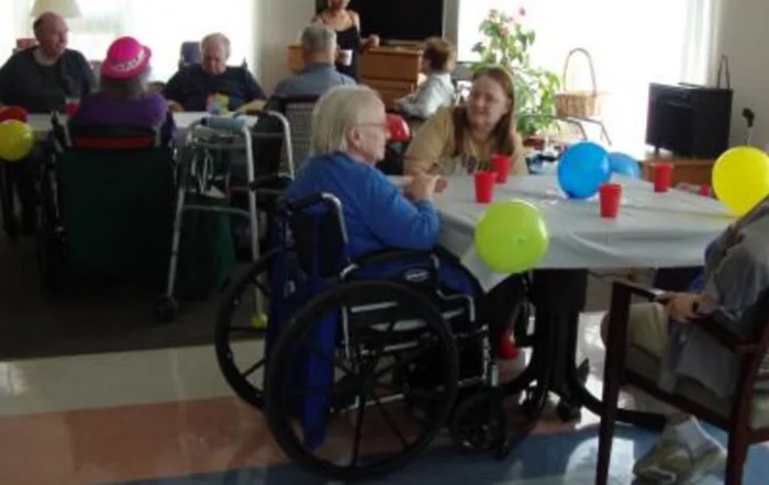 A social gathering at a nursing home with residents and staff interacting around a table decorated with colorful balloons.