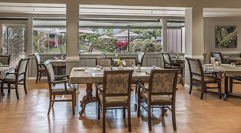 A cozy restaurant dining area with elegantly set tables and chairs, overlooking a garden through large windows.