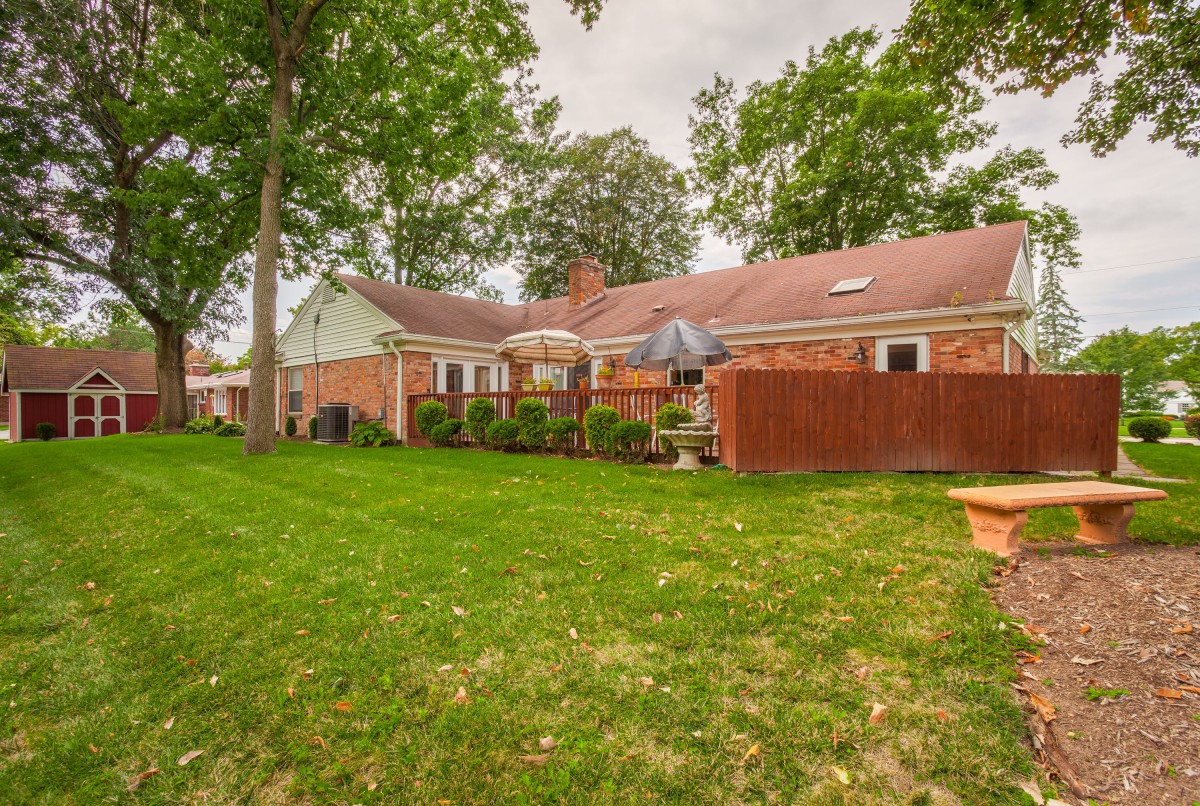 A suburban backyard with a lawn, trees, and outdoor seating area next to a brick house.