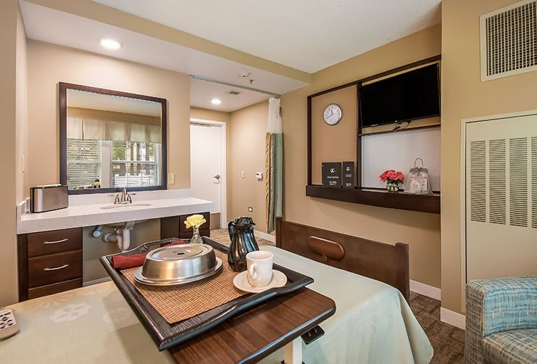 A modern hospital room featuring a dining table with a food tray, television, and a sink.