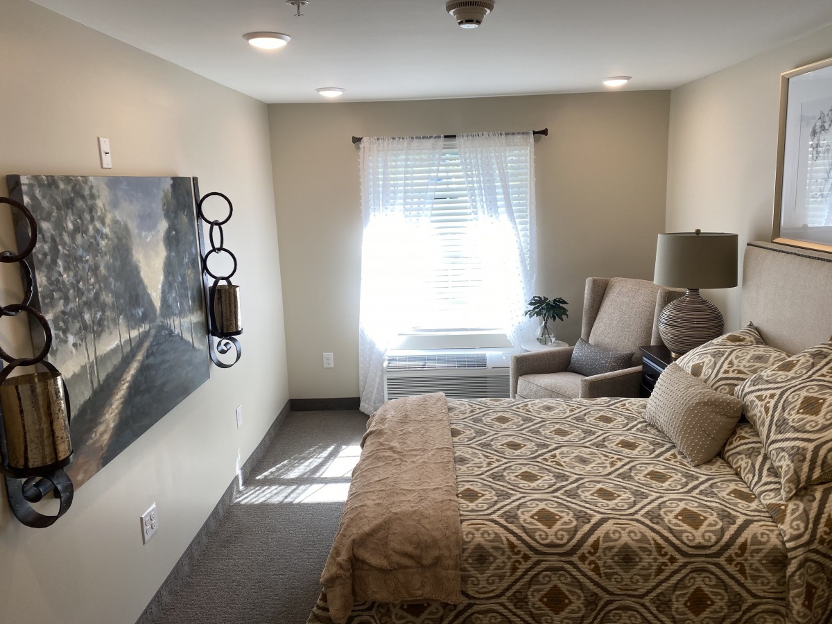 A cozy bedroom with a neatly made bed, armchair, and bright natural light coming through the window.