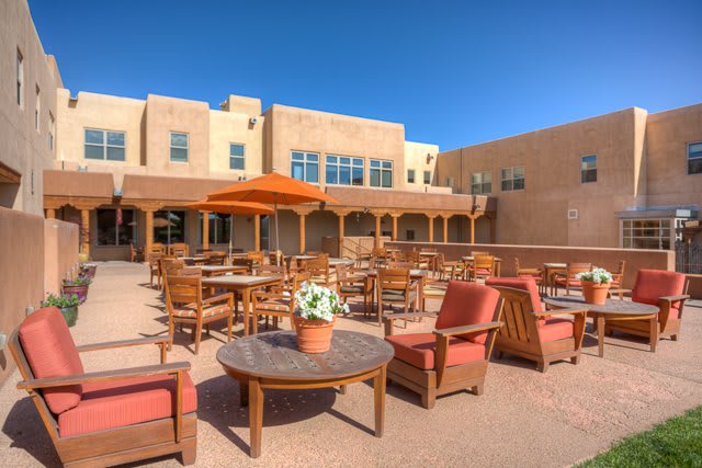 A sunny patio with wooden furniture and an umbrella, set against a building with desert-style architecture.