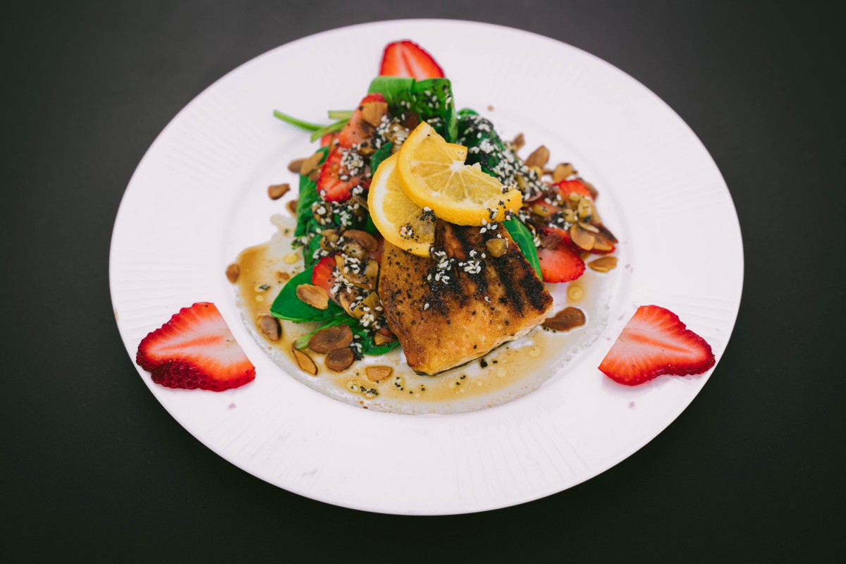 A gourmet dish featuring salmon served with strawberries, nuts, spinach, and a lemon garnish.