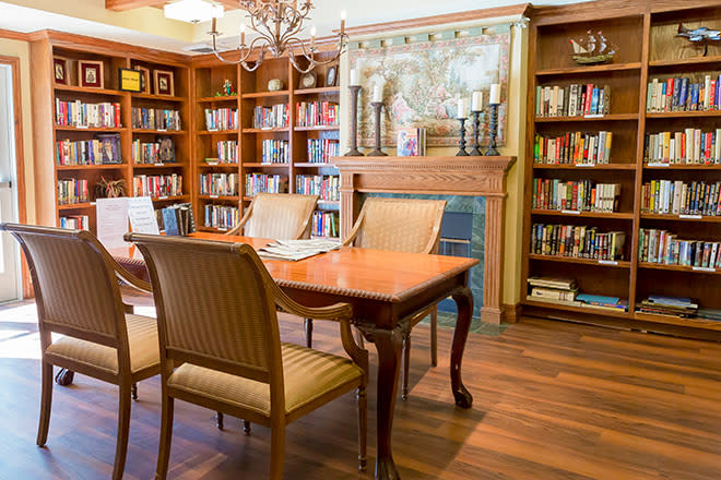 A cozy library room featuring a wooden table and chairs, surrounded by bookshelves and a fireplace.