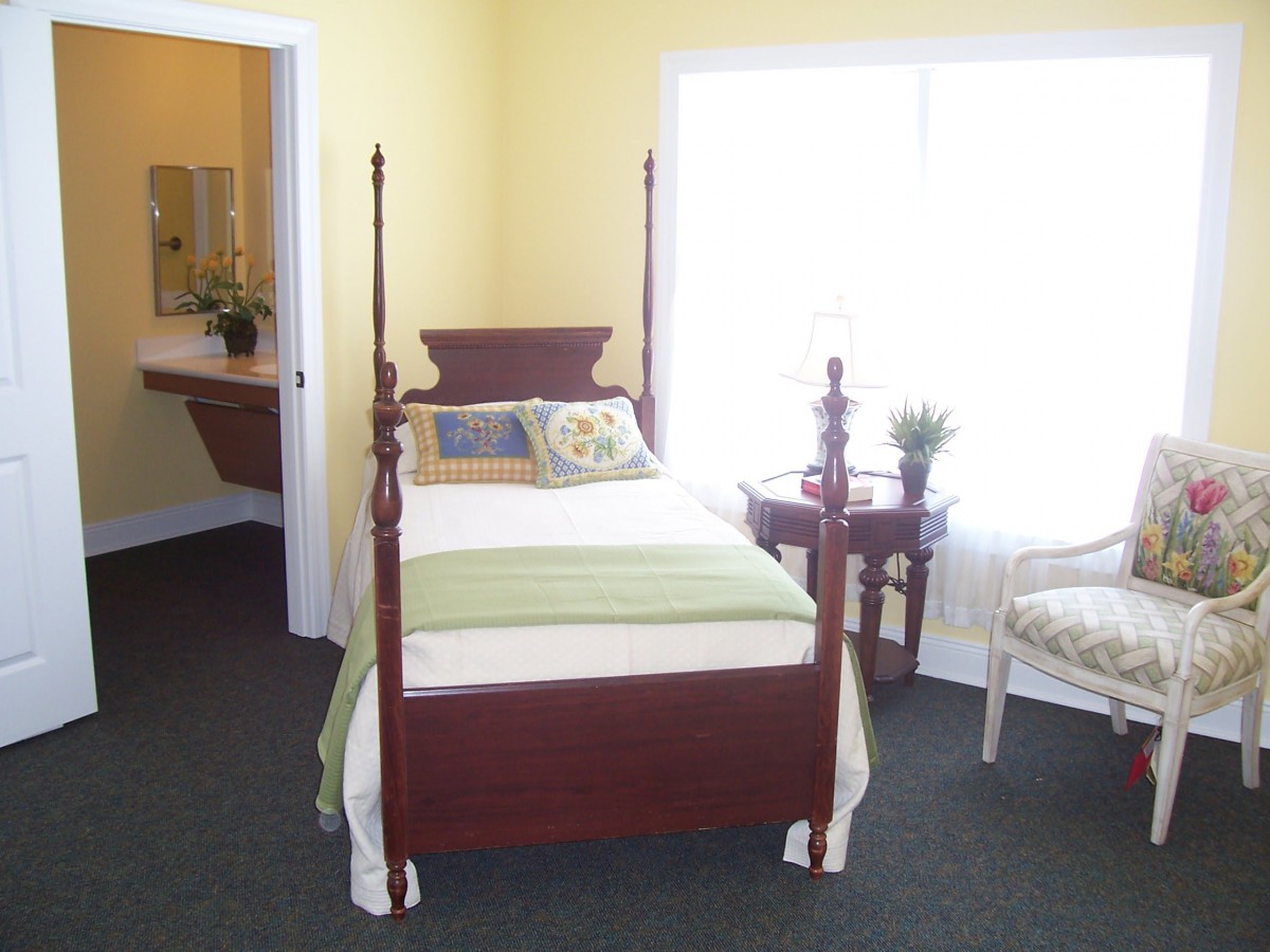 A cozy bedroom featuring a four-poster bed, a side table, and an upholstered chair, with natural light streaming in through the window.