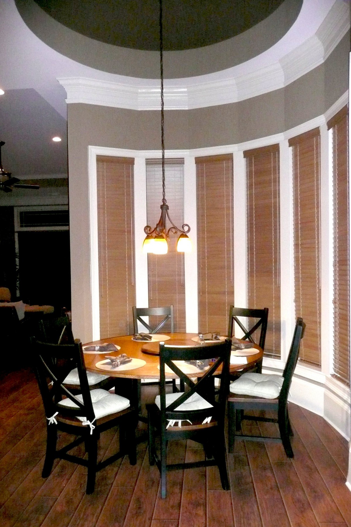 A cozy dining area with a round table, four chairs, and a pendant light fixture, surrounded by tall windows with blinds.