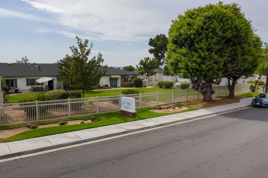 A suburban street with a fenced yard, trees, and a sign in front of residential buildings under a cloudy sky.