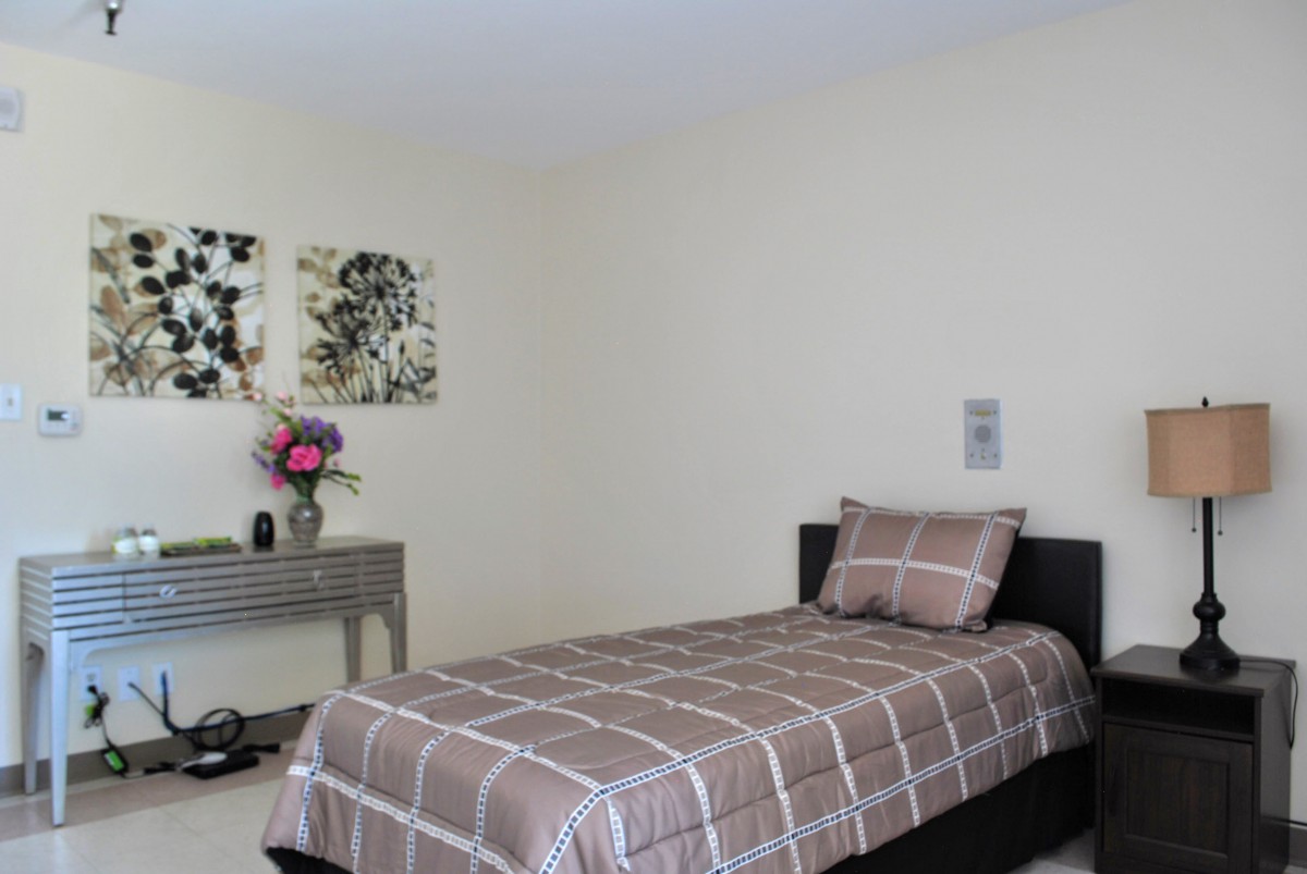 A minimalist bedroom featuring a single bed with brown plaid bedding, a decorative table with a vase of flowers, and artwork on the wall.