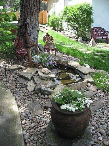 A serene backyard garden with a small pond surrounded by stones, lush greenery, and comfortable chairs for relaxation.