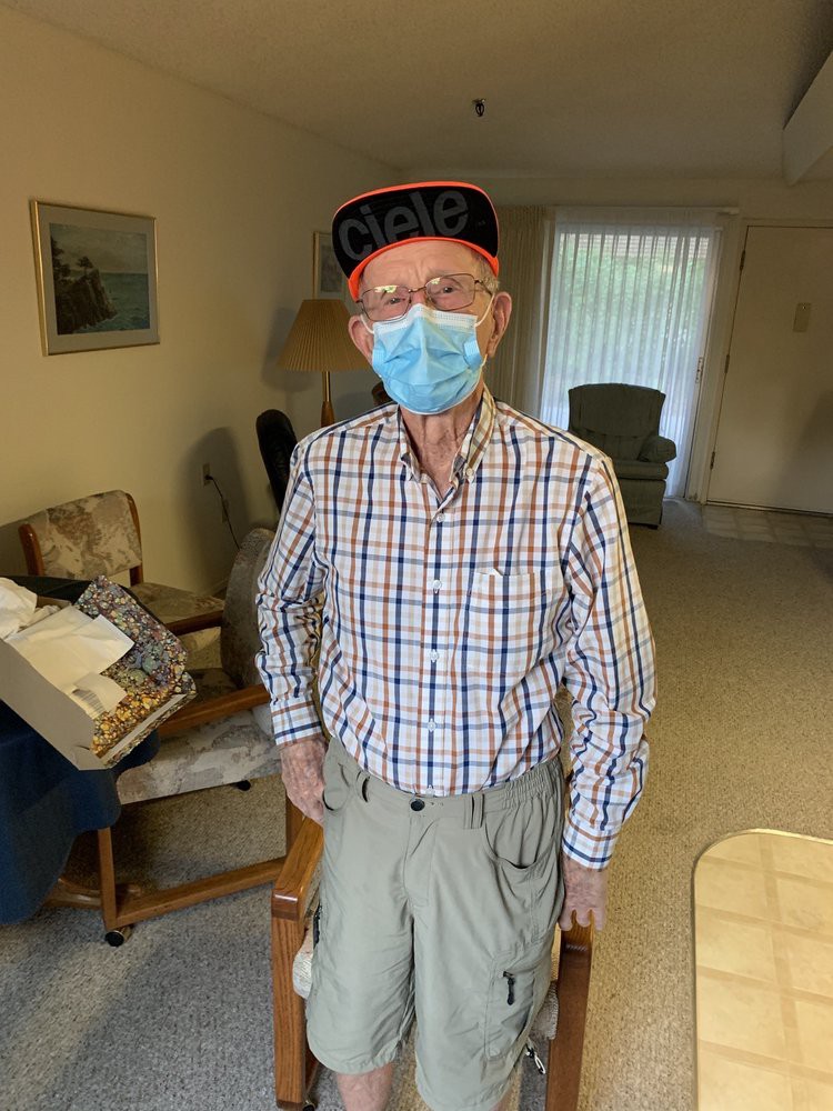 An elderly person wearing a face mask and hat stands in a living room.