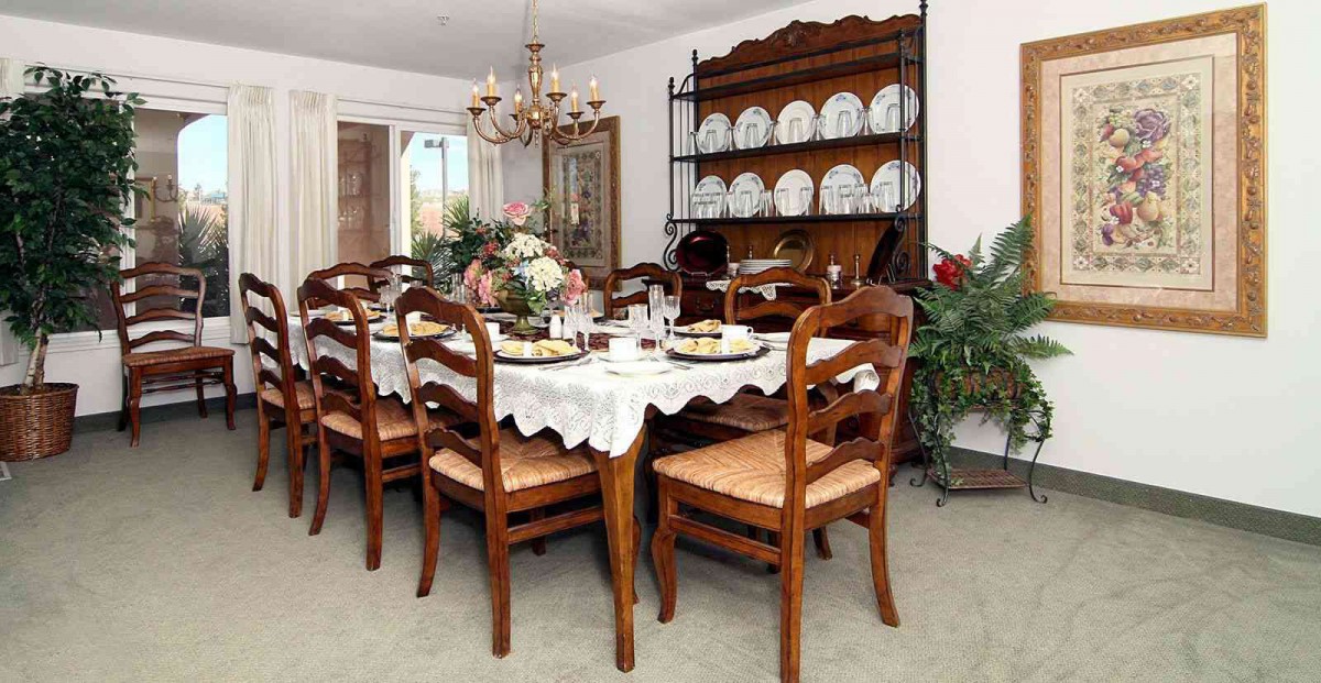A traditional dining room with a large wooden table set for a meal, surrounded by matching chairs. A chandelier hangs overhead, and a china cabinet stands against the wall displaying dinnerware.
