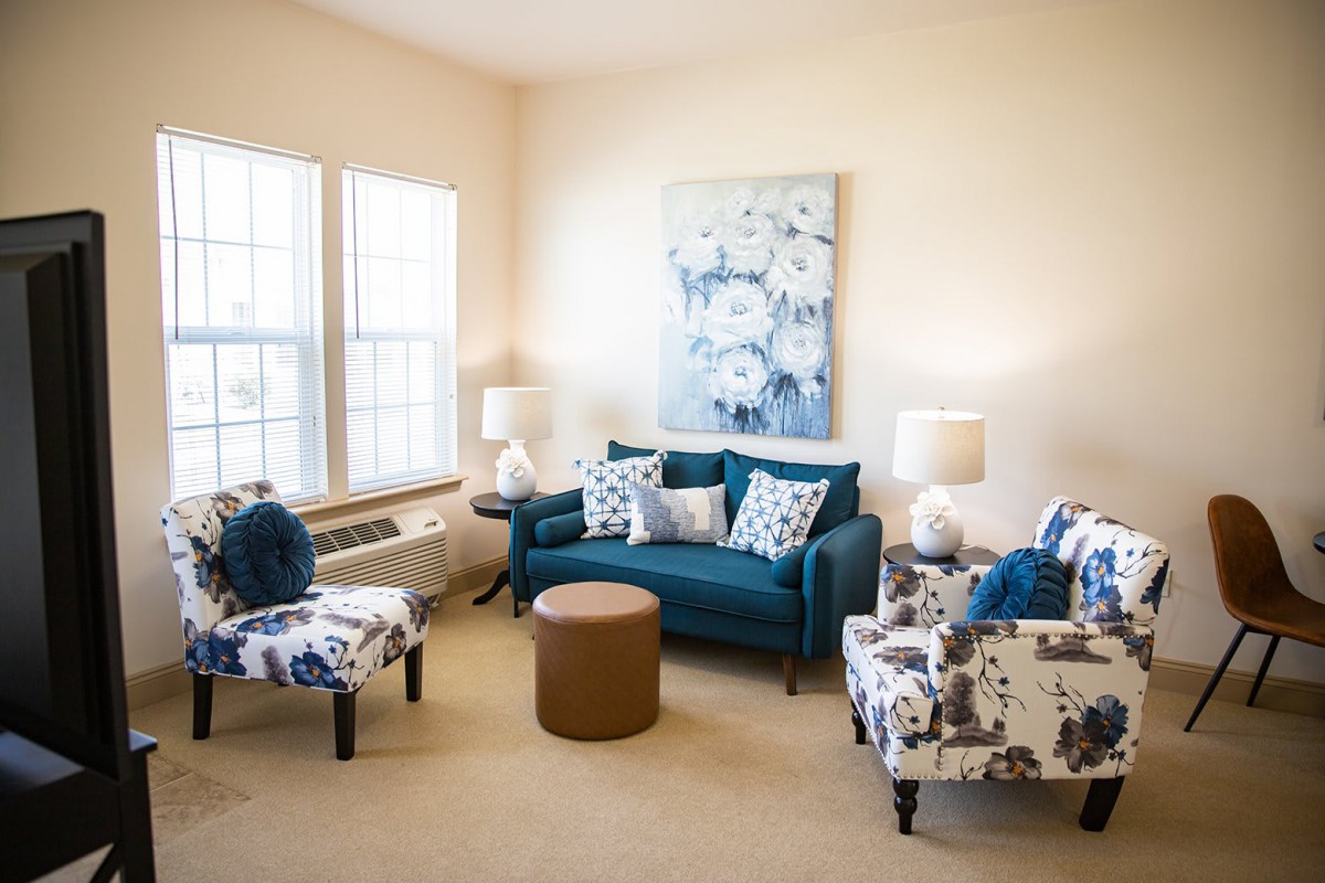 A cozy and well-lit living room featuring a teal sofa, patterned armchairs, an ottoman, and decorative artwork on the wall.