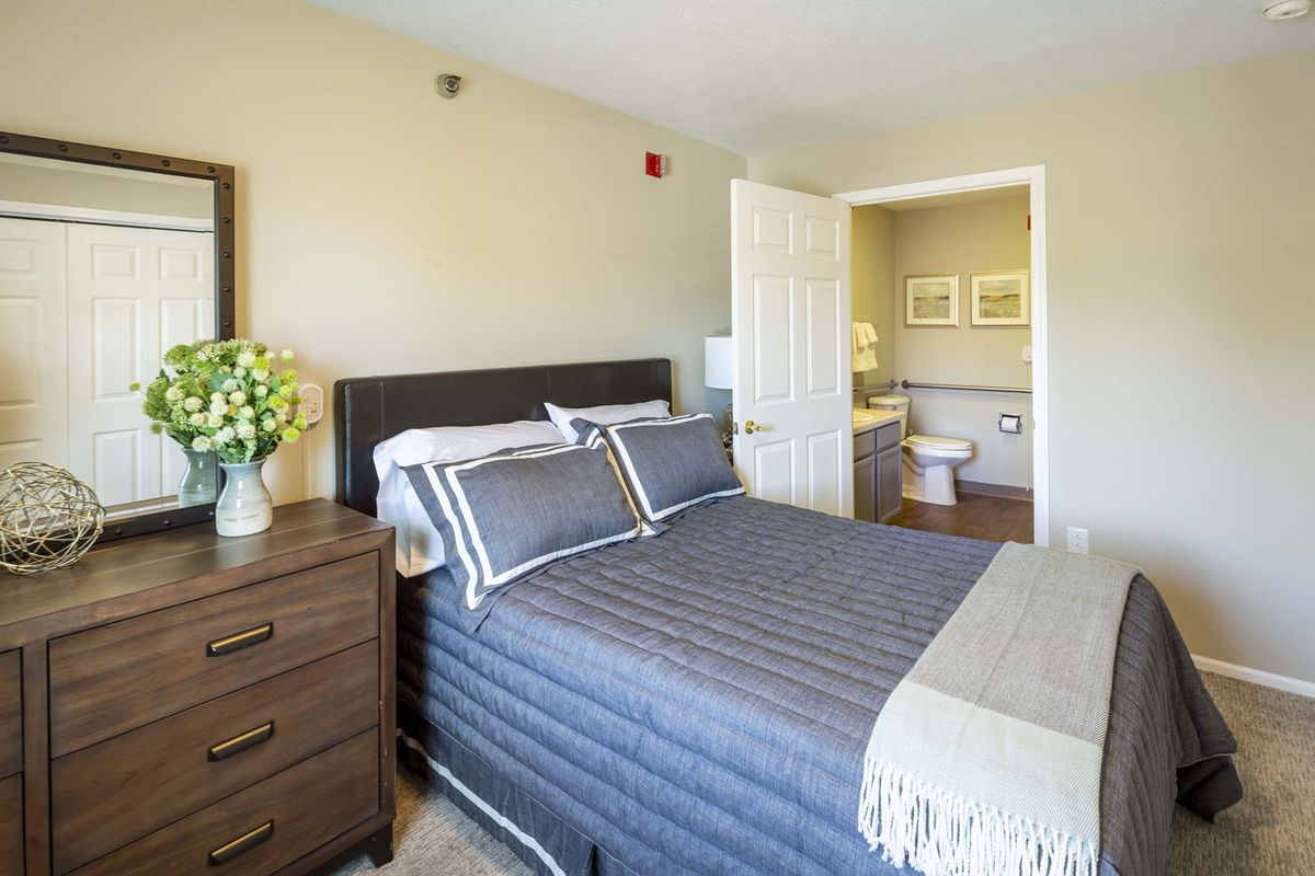 A cozy bedroom with a neatly made bed, nightstand with flowers, and an open door leading to the bathroom.