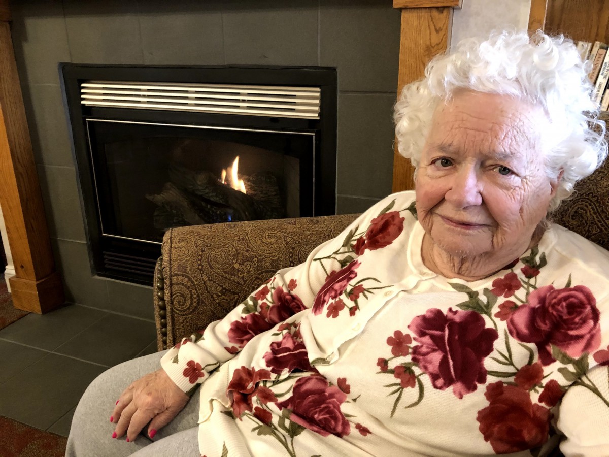 An elderly woman sitting comfortably by a fireplace, wearing a floral-patterned shirt.