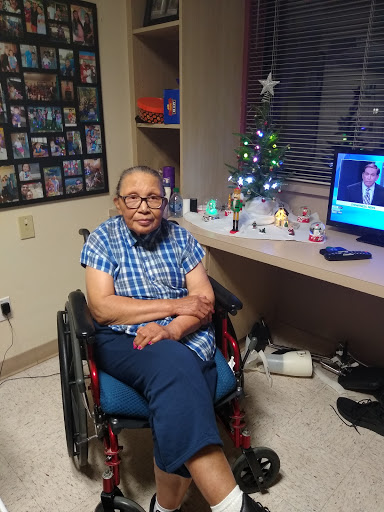 An elderly person sitting in a wheelchair, surrounded by holiday decorations and watching TV.