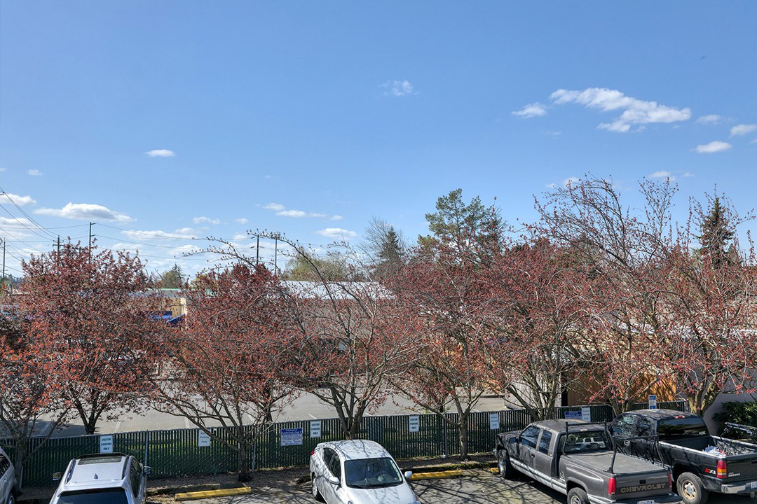 A parking lot with several cars under a clear blue sky and scattered clouds, surrounded by trees and buildings.