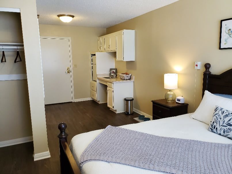 A cozy bedroom with a neatly made bed, bedside lamp, and kitchenette in the background.