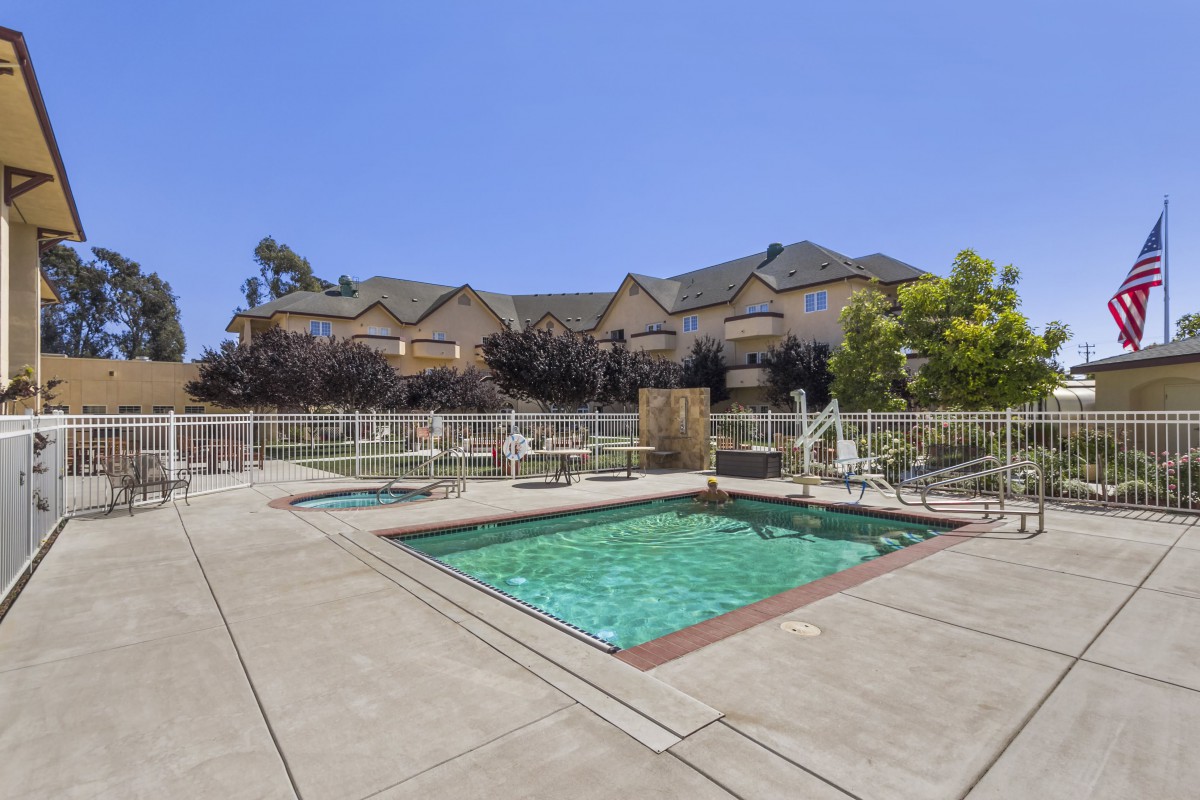 A residential outdoor area featuring a swimming pool and hot tub surrounded by a patio and a fence, with houses in the background under a clear blue sky.