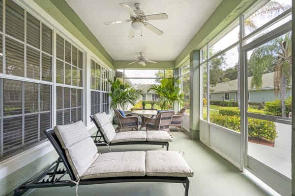 A bright sunroom featuring comfortable lounge chairs and a table, surrounded by large windows and lush plants.