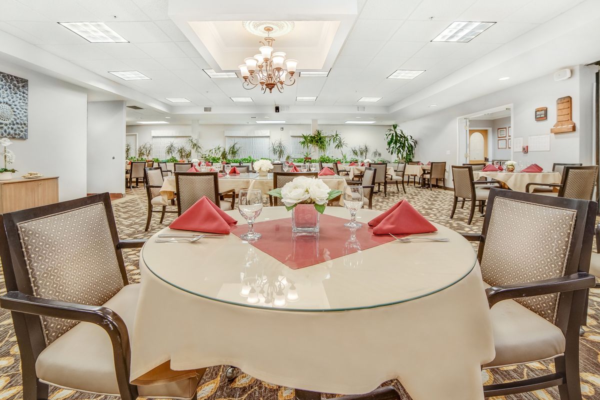 An elegant dining room with neatly arranged tables and chairs, featuring red napkins and floral centerpieces.