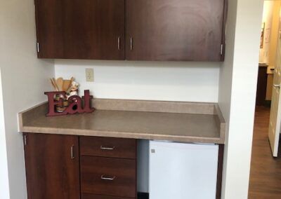 A small kitchenette with wooden cabinets and a countertop, featuring a mini fridge and decorative plants.
