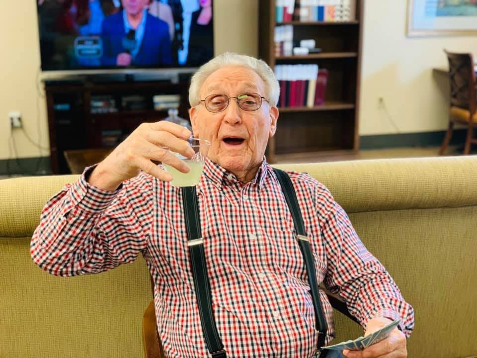 An elderly man wearing glasses and a plaid shirt is sitting indoors, holding up a drink and smiling.