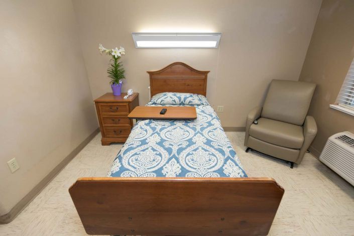 A hospital room with a single bed, nightstand with flowers, and a chair.