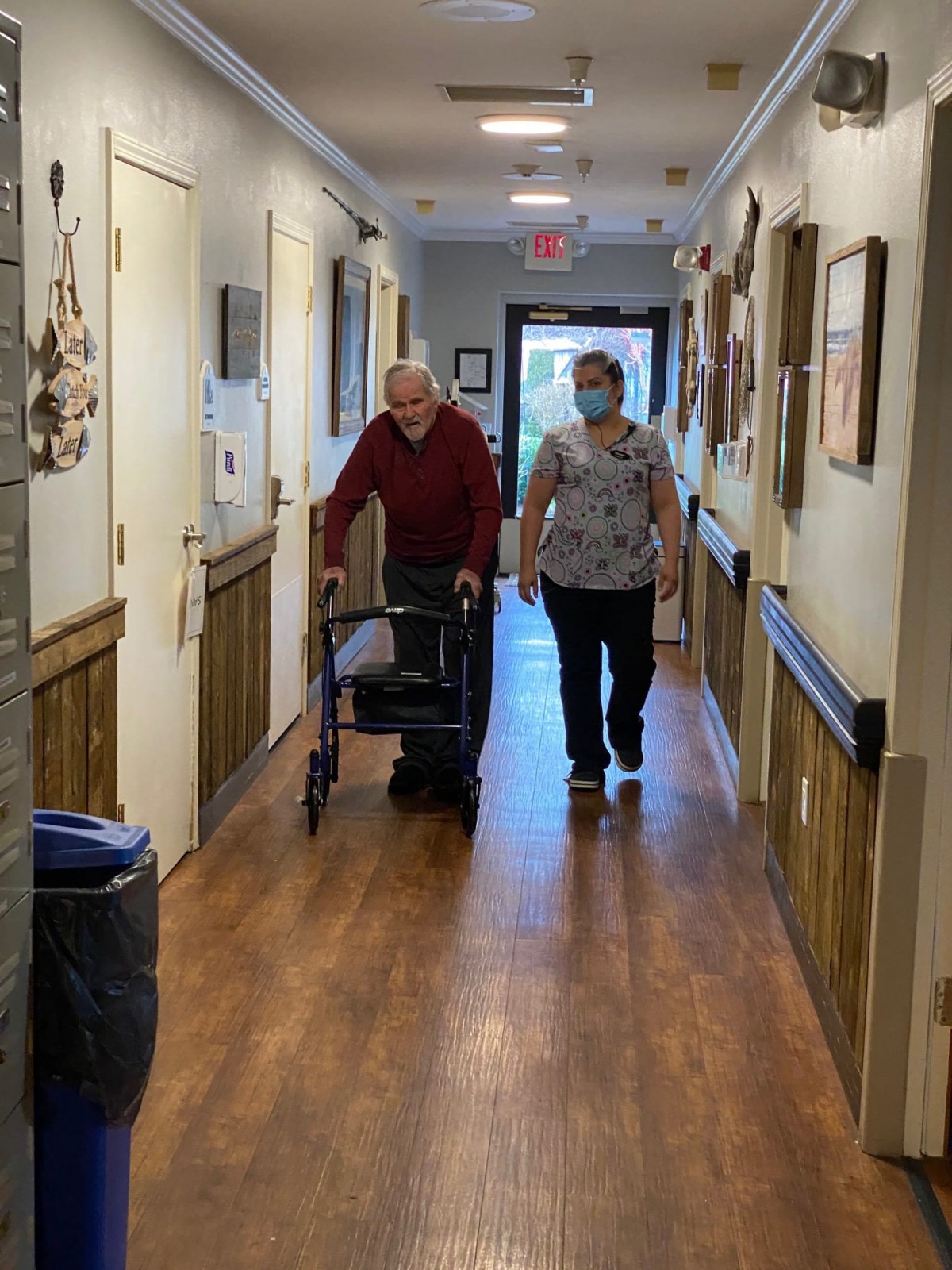 An elderly person using a walker is accompanied by a caregiver in a hallway of an assisted living facility.