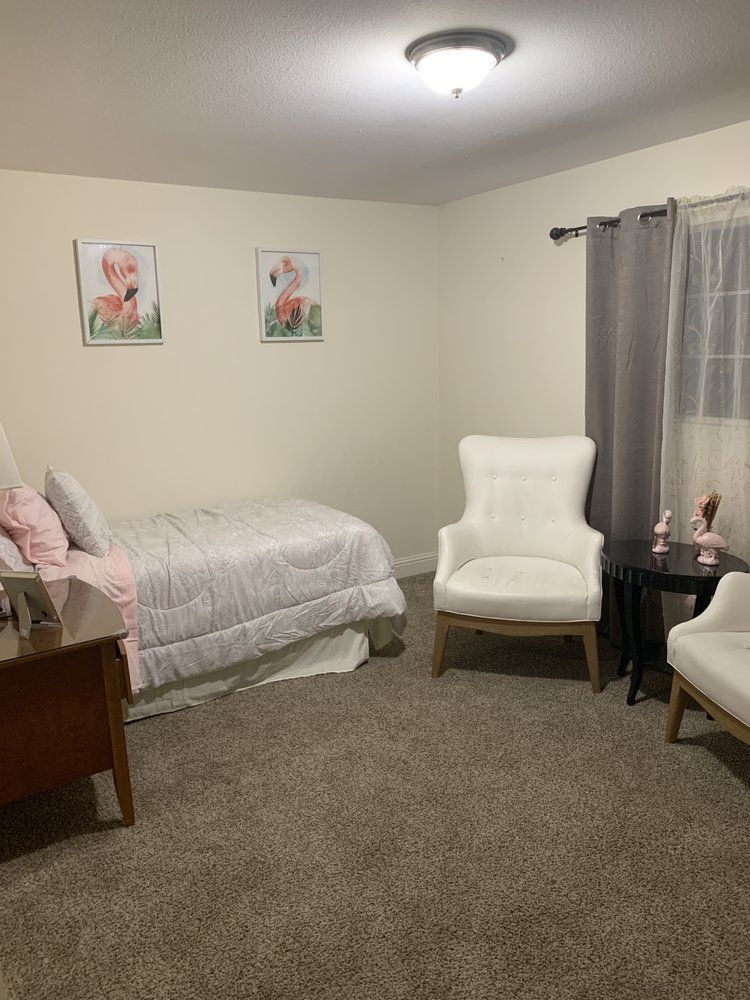 A cozy bedroom featuring a bed, two white chairs, a small table, and wall art.