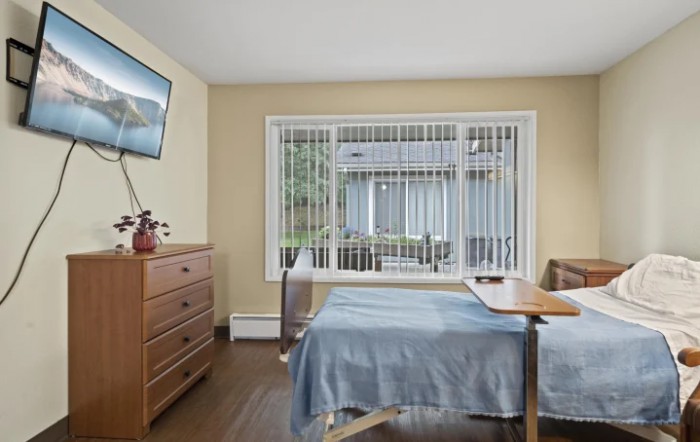 A cozy bedroom featuring a single bed with a blue cover, a dresser, and a mounted television. The room is well-lit by natural light coming through the large window.