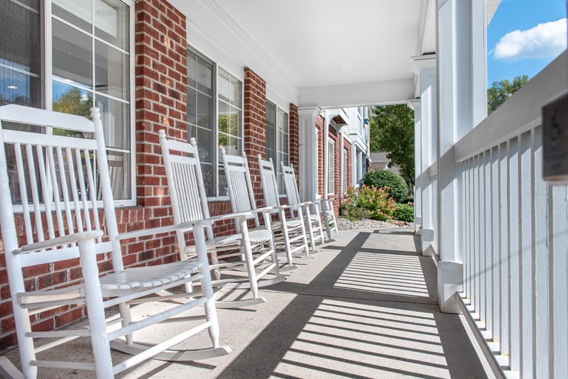A sunny porch lined with white rocking chairs against a brick wall.
