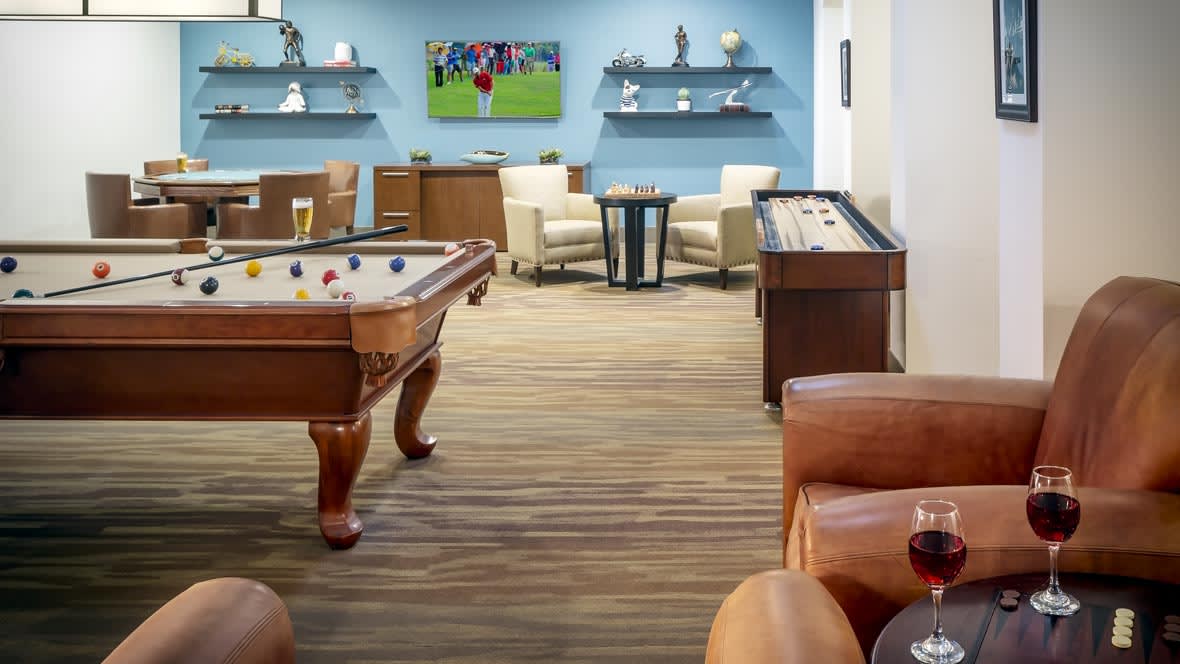 A modern game room featuring a pool table, shuffleboard, comfortable seating, and a television displaying a sports event.