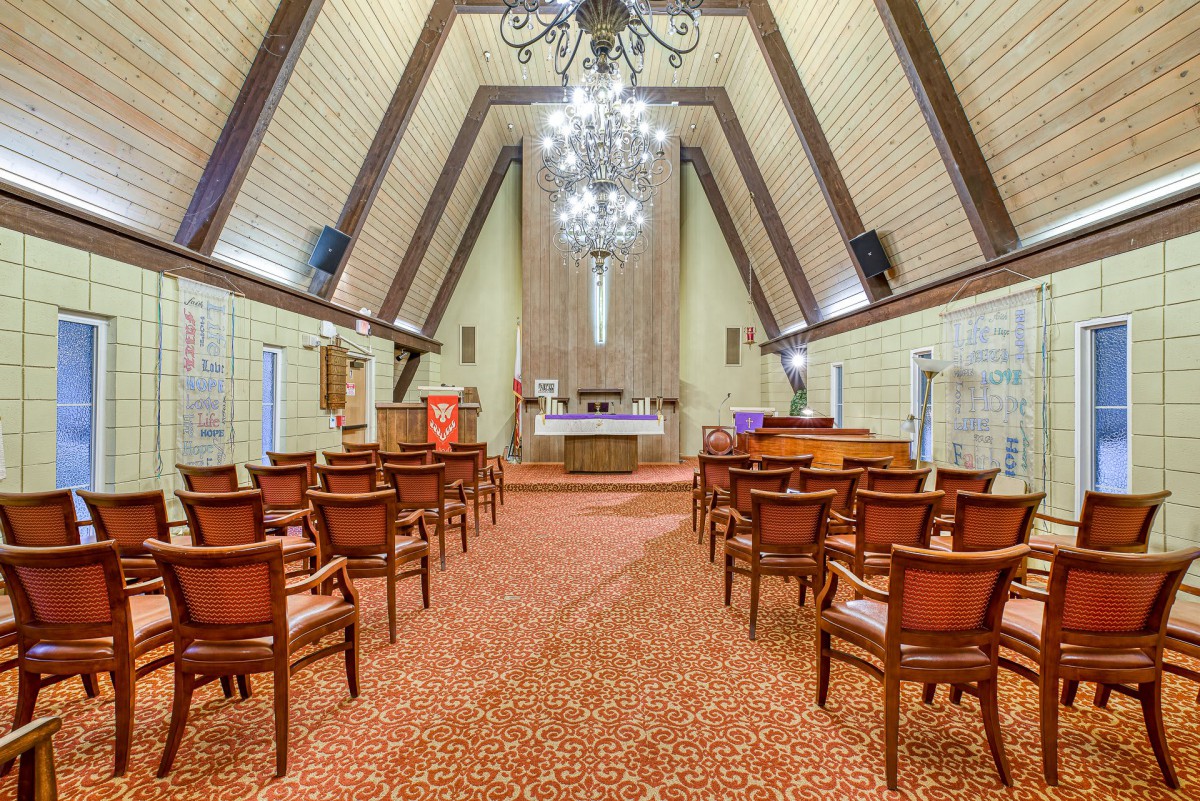 An elegant chapel interior with a central aisle, wooden chairs, and a prominent chandelier.