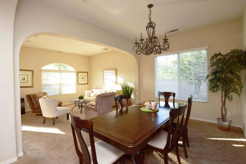 A dining area connected to a living room, featuring a wooden table and chairs under an ornate chandelier, with large windows providing natural light.