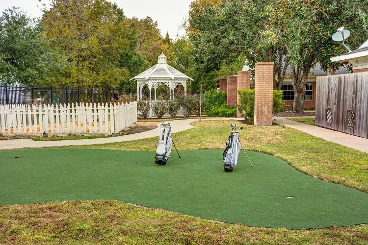 A serene outdoor setting featuring a small putting green with two golf bags, surrounded by trees and a gazebo.