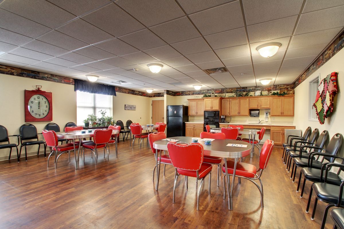 A spacious community dining area with red chairs and wooden tables, featuring a kitchen setup with cabinets and appliances.