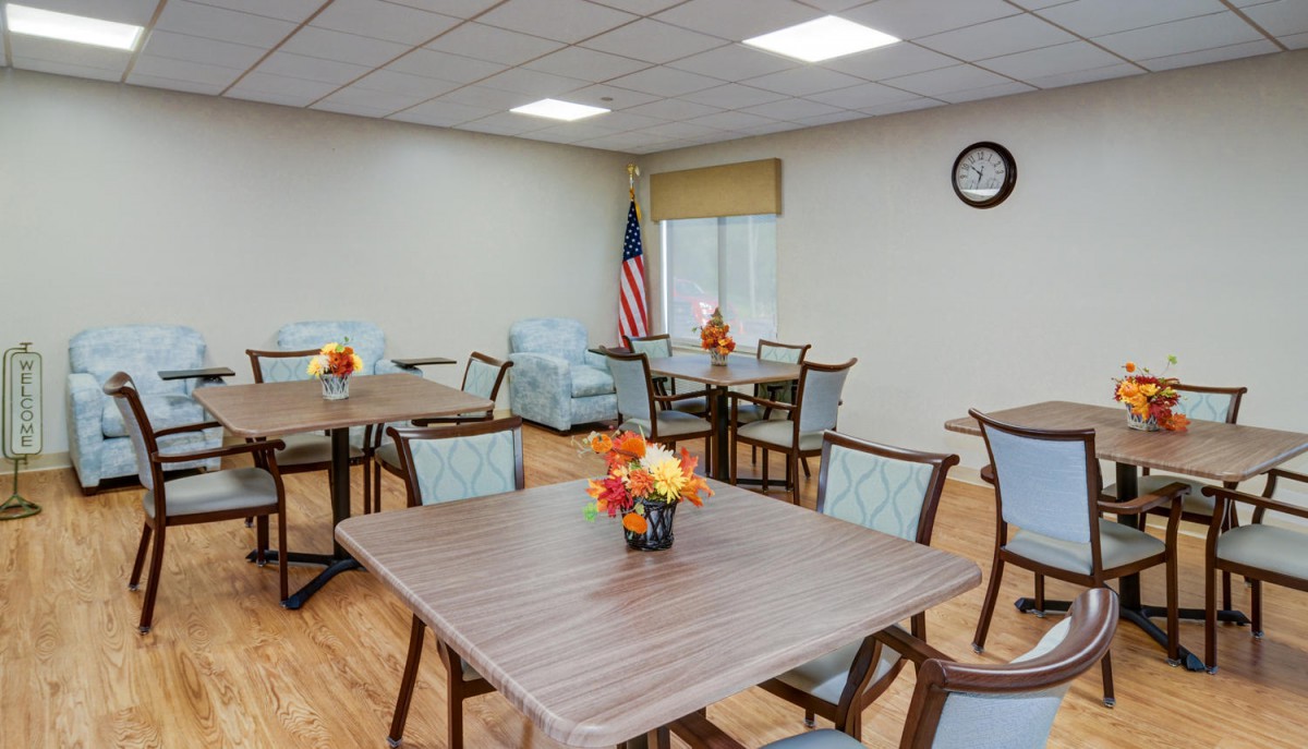 An empty dining room with tables and chairs, featuring floral centerpieces and an American flag in the corner.