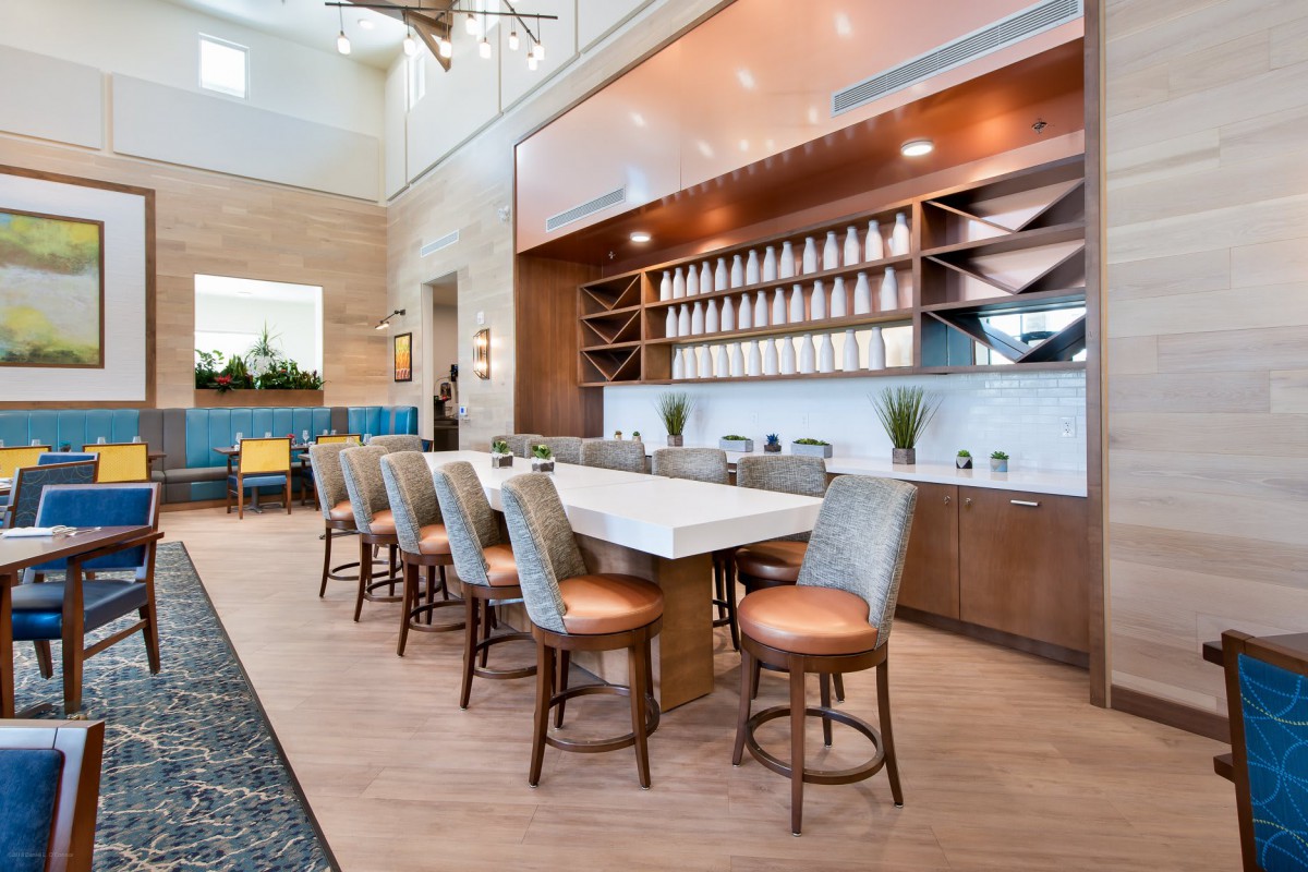 A modern kitchen interior featuring a long bar counter with upholstered chairs and open shelving.